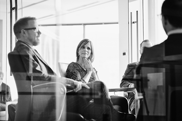 Black and white image of a group of people seated in a modern office setting. A woman is in the center, appearing thoughtful, with three others partially visible around her. Glass walls add a reflective, contemporary ambiance.