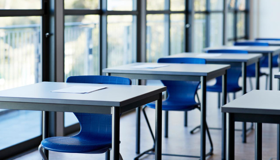 Empty classroom with rows of blue chairs and gray desks, each with a sheet of paper on top. Large windows line the left side, allowing natural light to brighten the room.