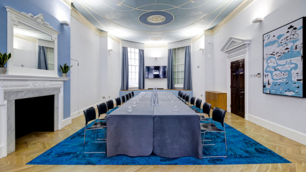 The Queen Elizabeth II room with a long, U-shaped table covered in a gray cloth, surrounded by black chairs. The room features a light blue and white color scheme, large windows, a decorative ceiling, a fireplace, and a piece of abstract art on the wall.