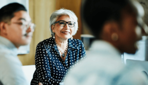 An older woman with glasses and gray hair smiles as she sits at a table in a meeting room. She wears a black and white polka dot blouse. Other people, slightly blurred, are in the foreground and background.