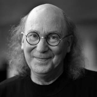 A person with long, light hair, round glasses, and a gentle smile is looking at the camera. The black-and-white image creates a soft and thoughtful atmosphere. The person is wearing a dark top.