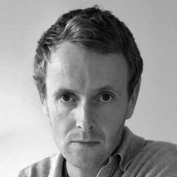 Black and white photo of a person with short, curly hair and a serious expression. They are wearing a collared shirt and looking directly at the camera. The background is plain and softly lit.