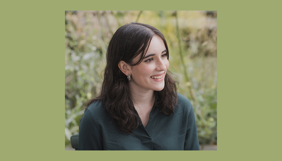 A woman with long brown hair is smiling and looking to the side. She is wearing a dark green blouse. The background features lush greenery. The image has a green border.
