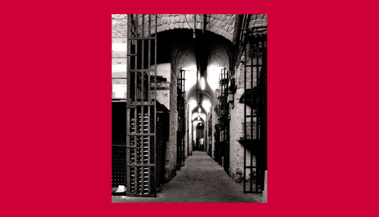A black and white photo of a narrow, dimly lit corridor with stone walls and archways. Steel gates with vertical bars line both sides, resembling prison cells. The scene is framed by a red border.