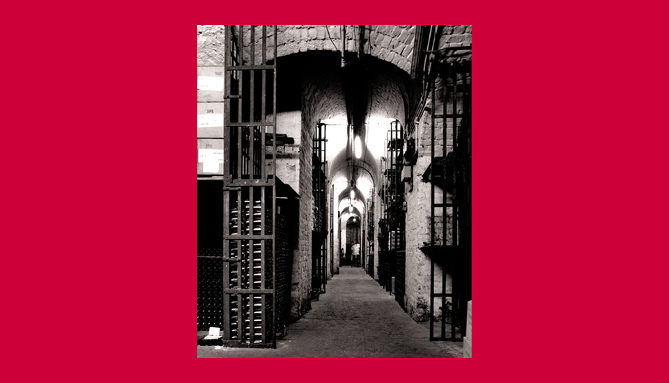 A black and white photo of a narrow, dimly lit corridor with stone walls and archways. Steel gates with vertical bars line both sides, resembling prison cells. The scene is framed by a red border.