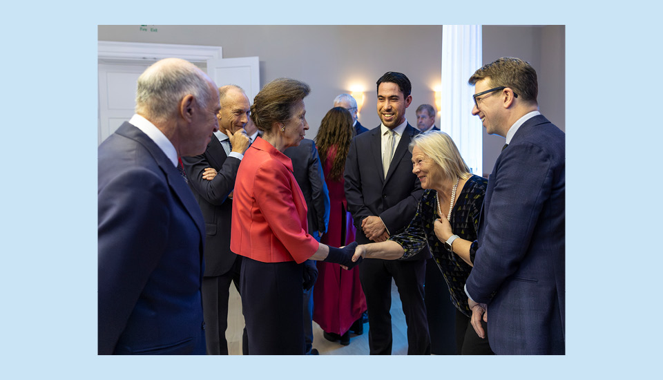 A woman in a red blazer is shaking hands with a smiling woman in a black dress. They are surrounded by several people in formal attire, engaged in conversation, in an elegant room.