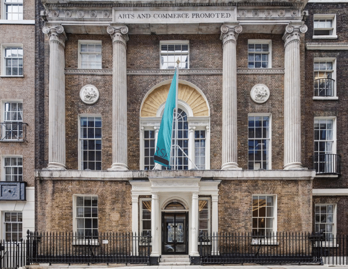 Historic building facade with tall columns and a large arched window, bearing a sign that reads "Arts and Commerce Promoted." A teal flag with "RSA" hangs from the entrance. The architecture features brickwork and classic design elements.