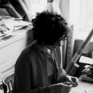 A person with curly hair and wearing a striped shirt is sitting by a cluttered desk, focused on their hands. The room is filled with scattered papers and art supplies. Sunlight streams in from a window to the right. The image is in black and white.