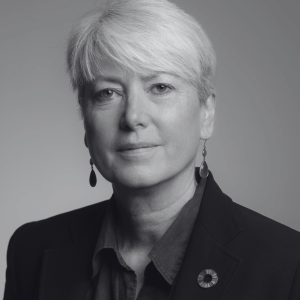 Black and white portrait of a person with short light hair wearing a dark blazer and drop earrings. The person looks directly at the camera with a neutral expression. The background is a smooth gray gradient.