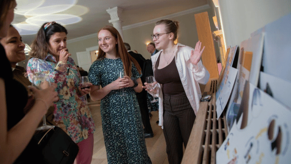 Four people stand in an art gallery, discussing prints displayed on the wall. They hold wine glasses and wear casual, colorful clothes. One person gestures with their hand while engaging with the others.