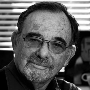 Elderly man with glasses and a beard, looking at the camera with a slight smile. Black and white photo with blinds in the background.