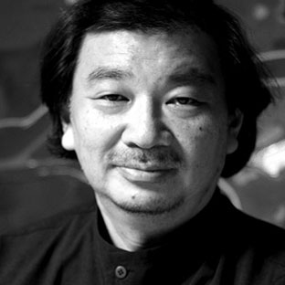 Black and white portrait of a person with medium-length hair and a mustache, wearing a dark top. The background is blurred and indistinct.