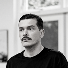 Black and white portrait of a man with a mustache, wearing a dark shirt. He is looking off to the side, standing indoors with blurred windows in the background.
