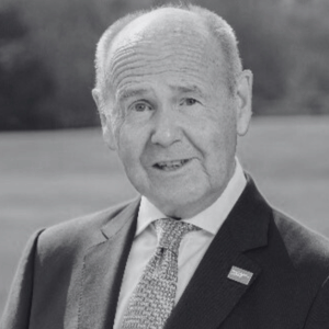 An elderly man in a suit and tie, wearing a pin on his lapel, smiles at the camera. The background is a blurred outdoor setting. The image is in black and white.