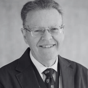 A smiling older man with glasses, wearing a suit and patterned tie. The image is in black and white.