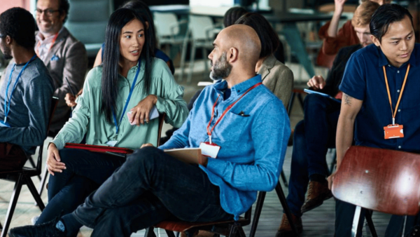 A group of people sitting on chairs in a casual office setting. A woman and a bald man in the foreground are engaged in conversation. Others are either talking or focused on different activities, all wearing name tags.