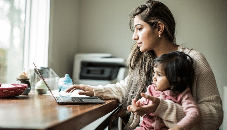A woman sits at a wooden table, working on a laptop, while holding a toddler on her lap. The woman looks focused on the screen. A window offers natural light, and there is a basket and a baby bottle on the table.