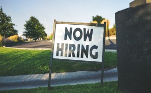 A sign reads "Now Hiring" in bold black letters on a white background, positioned on a grassy area near a road. Trees and a clear sky are visible in the background, suggesting a sunny day.