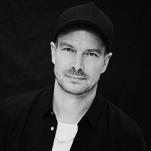 A person wearing a dark shirt, white top, and cap poses against a black background. They have a neutral expression and are facing the camera.
