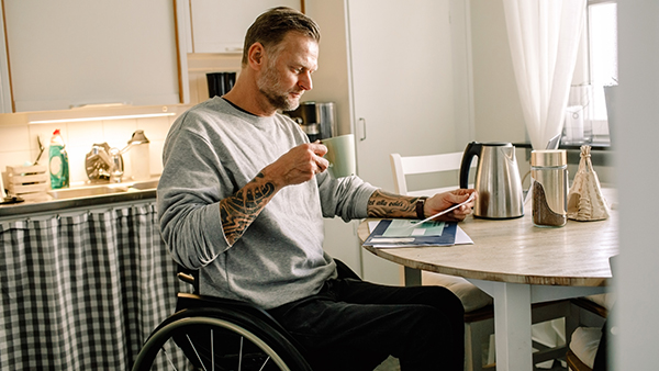 A man in a wheelchair sits at a kitchen table, holding a cup and reading a paper. He has tattoos on his arms and wears a gray sweatshirt. The kitchen includes a countertop, cleaning supplies, a kettle, and a window with curtains.