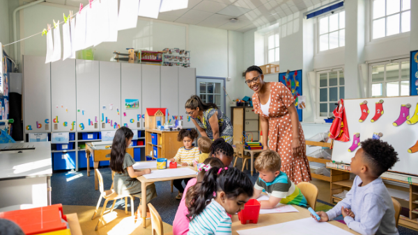 A vibrant classroom with young children seated at tables, engaged in activities. Two teachers are interacting with the students, one smiling at a child. The room is decorated with art supplies, a clothesline with papers, and colorful wall art.