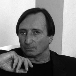 Black and white photo of a man with medium-length dark hair, wearing a dark jacket and shirt. He is resting his chin on his hand with a thoughtful expression.