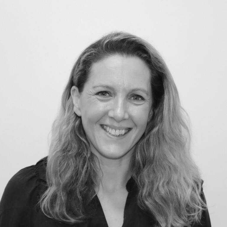 Black and white image of Vicky Kington smiling. She has long, wavy hair and are wearing a dark top. The background is plain.