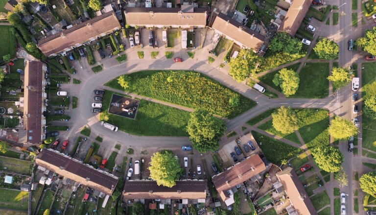 Aerial view of a residential area with houses arranged in a semicircle around a central green space with trees. Cars are parked along the roadways, and pathways intersect the grassy area. The setting is surrounded by lush greenery.