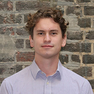 A person with curly brown hair wearing a light-colored, checkered shirt stands against a textured brick wall, looking at the camera with a neutral expression.