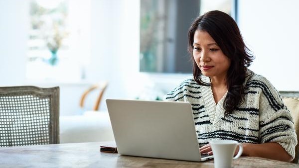 A person sits at a wooden table using a laptop, appearing focused. They wear a black and white striped sweater. A smartphone and a white mug are on the table. The background is bright and softly blurred.