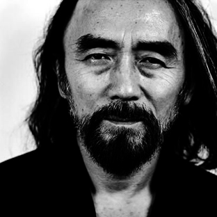 Black and white portrait of a man with long hair, a beard, and mustache. He is wearing a dark shirt and looking directly at the camera with a slight smile. The background is plain and out of focus.