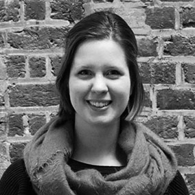 A person with short hair smiles in front of a brick wall. They are wearing a scarf and a dark top. The photo is in black and white.