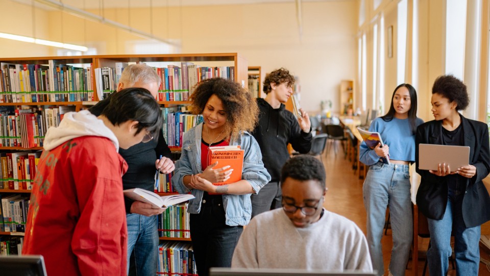A group of people in a library. Some are standing near bookshelves, holding and discussing books, while others are using laptops and tablets. The room is well-lit, with rows of books and tables.