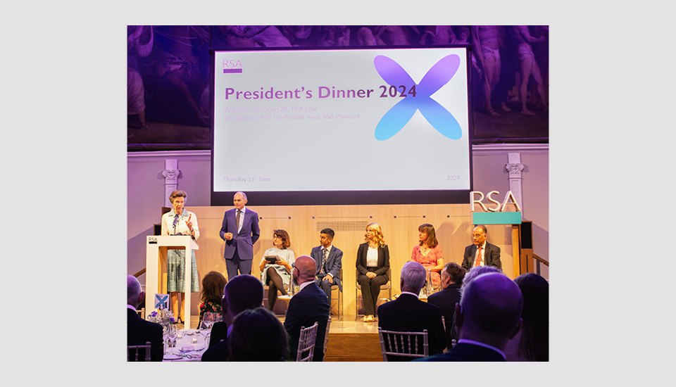 A group of people sits on a stage under a large screen that reads "President's Dinner 2024" at the RSA venue. A woman and a man stand at a podium. The background features an ornate mural.