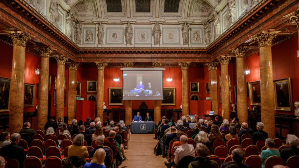 A large group of people sits in an ornate hall with red walls and golden columns, watching a presentation on a screen at the front. The ceiling is intricately decorated, and framed paintings line the walls.