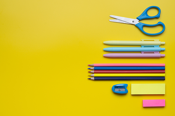 Colorful school supplies arranged on a yellow background. Items include a pair of blue-handled scissors, three pens in green, blue, and pink, three colored pencils in red, pink, and blue, a green eraser, and a blue sharpener.