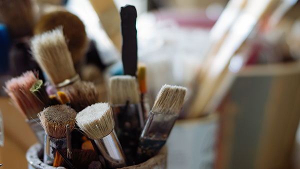 A close-up of assorted paintbrushes with varying sizes and shapes, some with frayed bristles, in containers. The background is blurred, highlighting the texture and details of the brushes.