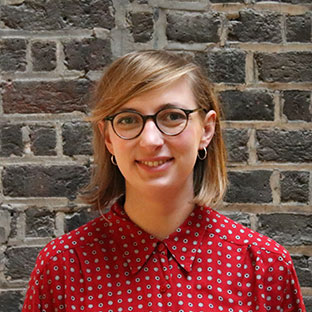 A person with shoulder-length hair and glasses smiling, wearing a red patterned shirt. They are standing in front of a brick wall.
