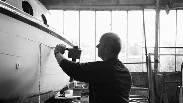 A person is shaping a wooden boat with a hand tool in a workshop. The room is lit by large windows in the background, casting light on the boat's hull. The black-and-white scene highlights the craftsmanship involved.