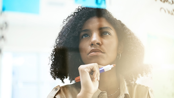 Woman with curly hair, holding a whiteboard marker, gazes thoughtfully at a semi-transparent surface covered with indistinct writing. She appears focused, with a light, blurred background.