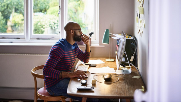 A person with a beard sits at a wooden desk in a bright room, working on a computer. They hold their glasses in one hand and have a focused expression. The desk is cluttered with papers, and a lamp is beside the monitor. Large windows are in the background.