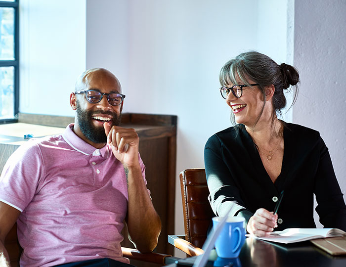 Two people sit at a table, smiling and engaging in conversation. The man on the left is in a pink shirt, while the woman on the right has glasses, a black top, and holds a pen over a notepad. Sunlight filters in through a nearby window.