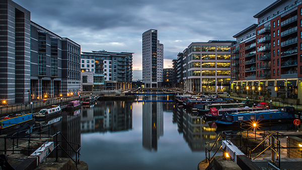 A calm, illuminated canal lined with modern high-rise buildings and moored boats at dusk. The sky is overcast, with reflections visible on the water.