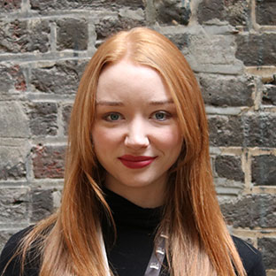 A person with long red hair and a black top stands in front of a brick wall, smiling softly at the camera.