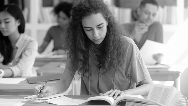 A black and white image of a young woman with curly hair sitting at a desk in a classroom. She is focused on writing in a notebook, with an open book in front of her. Other students are also sitting, reading, and writing in the background.