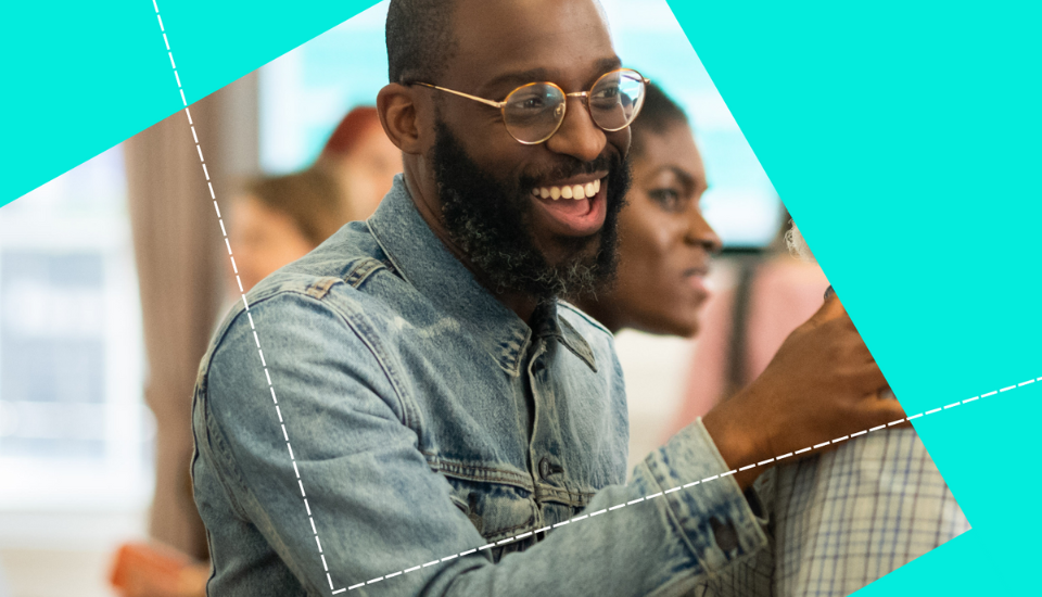 A man with a beard and glasses is smiling and wearing a denim jacket. He is interacting with another person in a lively setting. The image has a creative overlay of teal shapes and dotted lines.