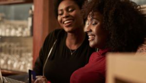 Two smiling women sit together indoors, one holding a credit card while looking at a laptop. Shelves with glasses and bottles are visible in the background.