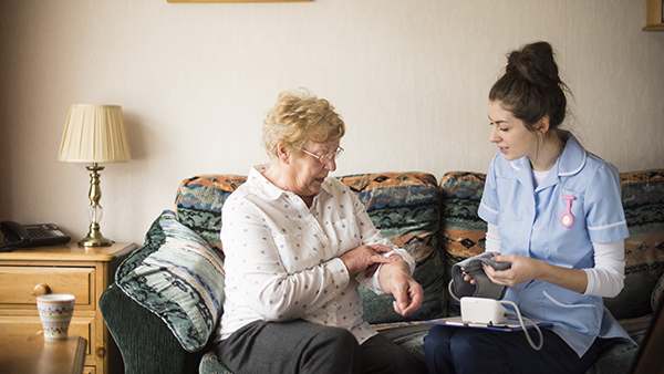 A nurse in blue uniform sits on a couch next to an elderly woman, checking her blood pressure. The woman holds out her arm with a cuff on it. A lamp and wooden side table with a mug are in the background.