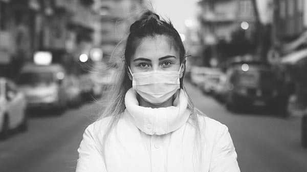 A woman wearing a face mask and a high-collared jacket stands on a street. The background is slightly blurred, showing parked cars and buildings on either side, creating an urban setting. The image is in black and white.
