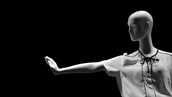 Mannequin in a white blouse with a black ribbon around the neckline, standing against a black background. Its right arm is extended to the side, creating a dramatic contrast with the dark backdrop.
